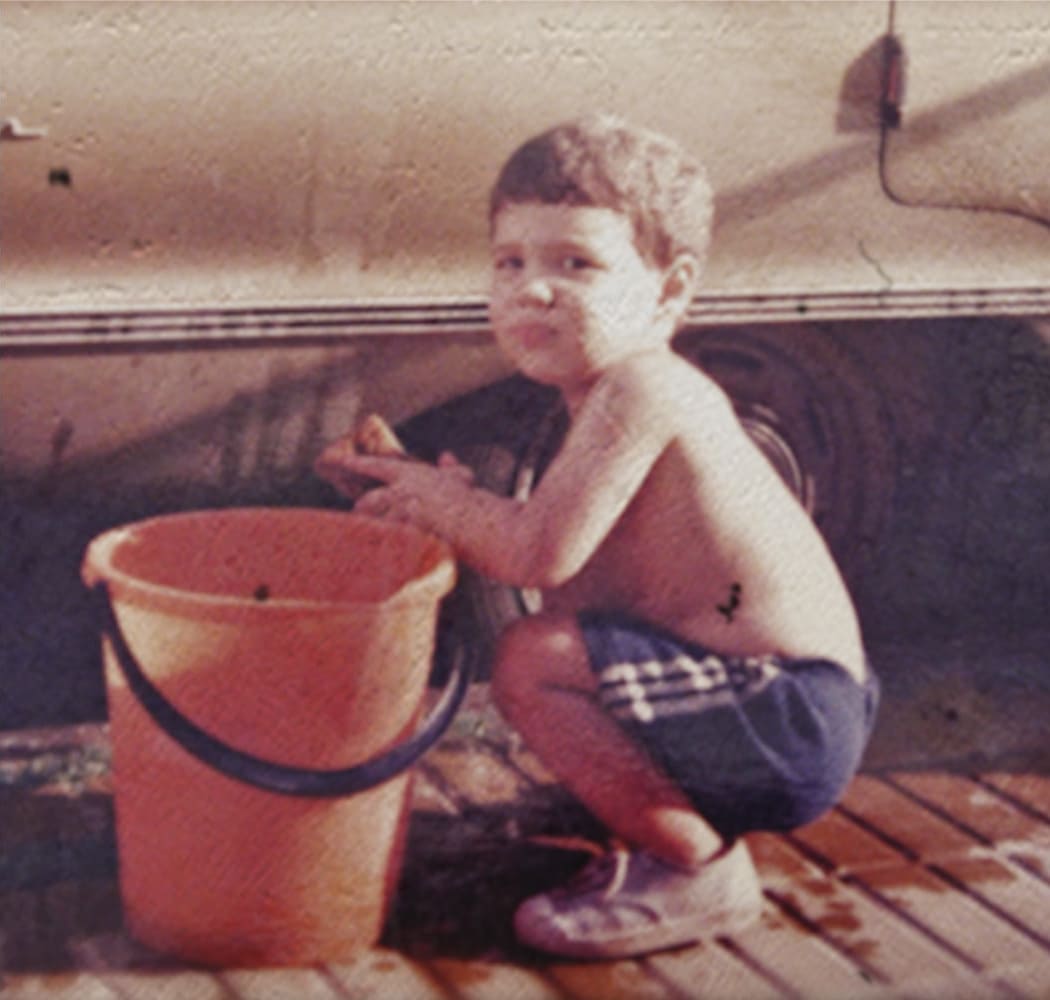 Nene agachado con un balde, al lado de la rueda de un auto.